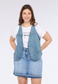 Džínová vesta v světlé modré barvě s bezramenným designem, dvěma předními kapsami a kontrastním prošíváním, vrstvená přes bílé tričko s potiskem a modrou sukni.