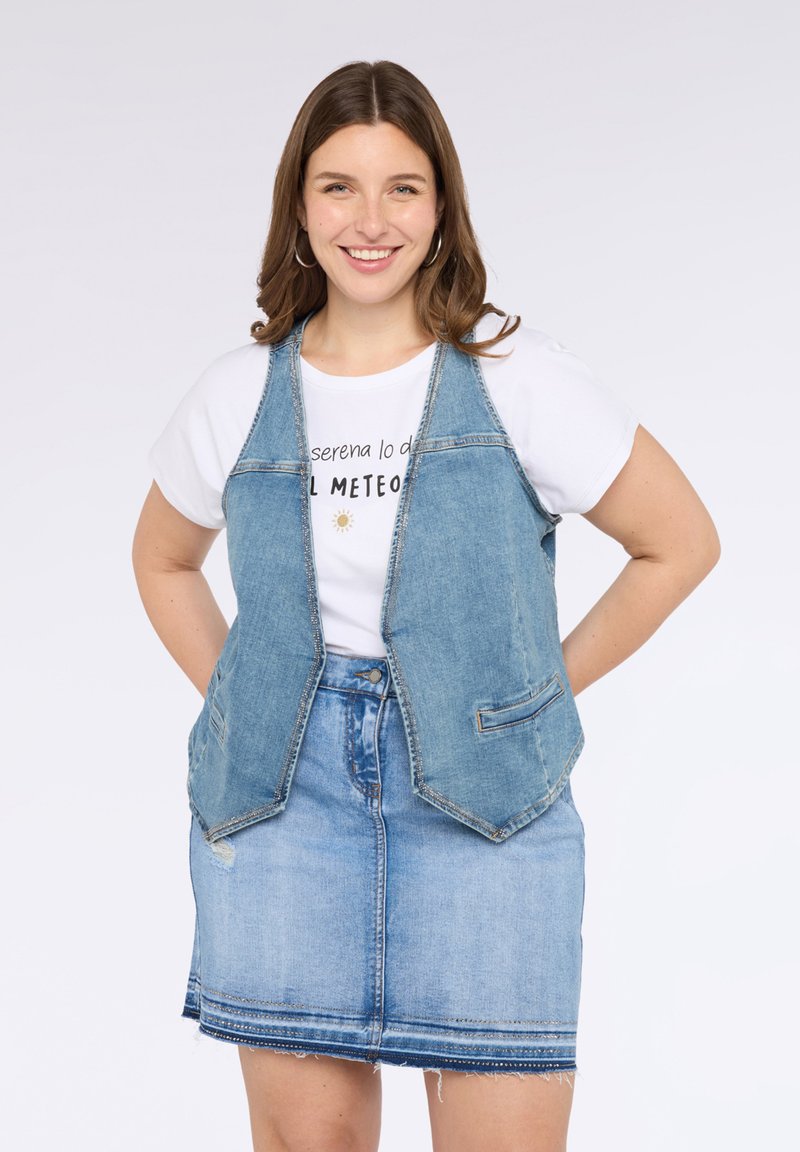 Džínová vesta v světlé modré barvě s bezramenným designem, dvěma předními kapsami a kontrastním prošíváním, vrstvená přes bílé tričko s potiskem a modrou sukni.
