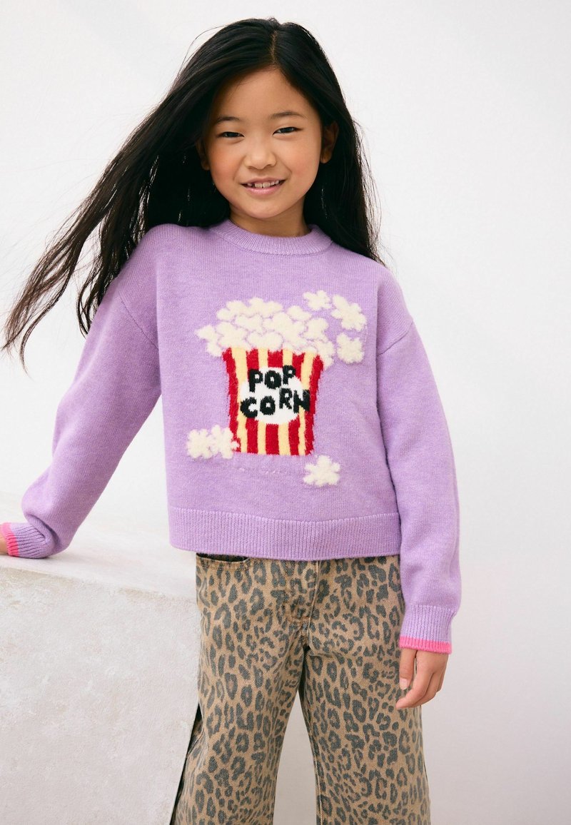 Pige med langt sort hår iført lilla sweater med popcorn-grafik og leopardmønstrede bukser, smilende mod en ensfarvet lys baggrund.
