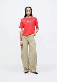 Nő fonott hajjal, piros Tommy Jeans pólót, bézs bő szárú nadrágot és fekete hegyes orrú cipőt viselve, egyszerű fehér háttér előtt áll.