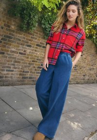 Rødternet button-up skjorte med lomme, parret med vide blå denim bukser, begge i en afslappet stil. Modellen står foran en murstensvæg.