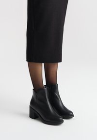 Czarne skórzane botki za kostkę o teksturowanym wykończeniu, z bocznym zamkiem błyskawicznym i masywnym obcasem, noszone z przezroczystymi czarnymi rajstopami i czarną spódnicą do kolan.