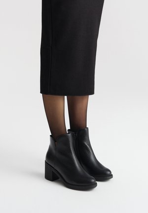 Czarne skórzane botki za kostkę o teksturowanym wykończeniu, z bocznym zamkiem błyskawicznym i masywnym obcasem, noszone z przezroczystymi czarnymi rajstopami i czarną spódnicą do kolan.