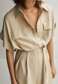 Beige napillinen mekko, jossa on kaulus, kaksi rintataskua, lyhyet hihat ja vyötärövyö. Sileä pinta ja rento mallisto.