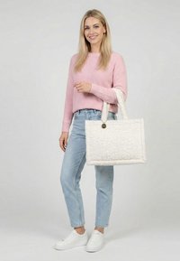 Nuori nainen, joka pukeutunut vaaleanpunaiseen neuleeseen ja vaaleisiin farkkuihin, pitää kädessään suurta valkoista teksturoitua kangaskassia, seisoo yksinkertaisen taustan edessä.