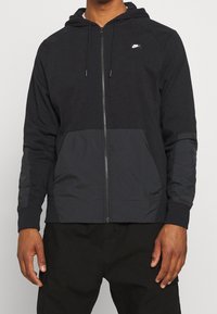 Svart zip-up hoodie med långa ärmar, med en texturerad kropp, kontrasterande släta paneler och en liten logotyp på bröstet.