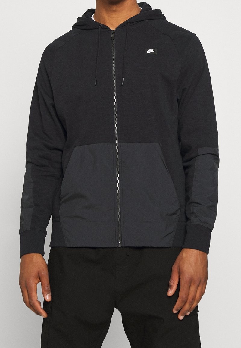 Svart zip-up hoodie med långa ärmar, med en texturerad kropp, kontrasterande släta paneler och en liten logotyp på bröstet.