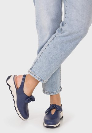 Pies con sandalias de cuña azul marino con detalles de lazo y suelas blancas gruesas, combinadas con vaqueros azul claro remangados.