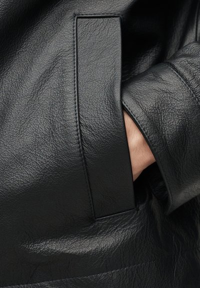 Χέρι μερικώς μέσα στην τσέπη ενός μαύρου δερμάτινου μπουφάν με υφή, που δείχνει λεπτομερή ραφή και υφή υλικού.
