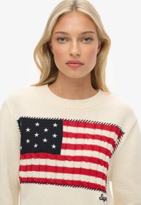 Sweter dzianinowy w kremowym kolorze z wyraźnym, teksturowanym wzorem amerykańskiej flagi; akcenty w kolorze czerwonym i niebieskim, z okrągłym dekoltem i widocznym szyciem.