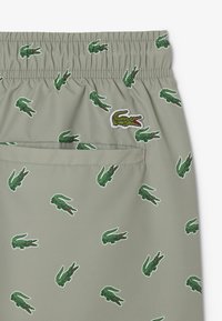 Lichtgrijze shorts met een elastische tailleband, voorzien van een herhaald groen krokodillenpatroon en een geborduurd krokodillenlogo aan de zijkant.
