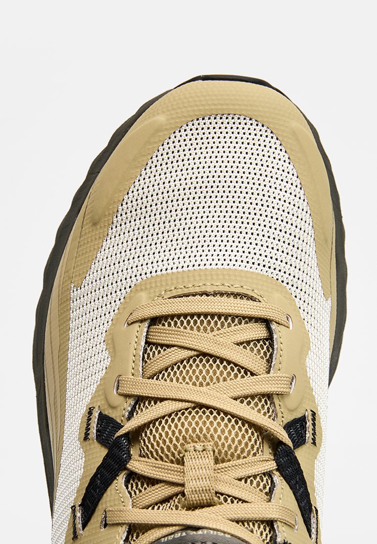 Zapatilla de malla beige y blanca con detalles en negro, cordones detallados y suela texturizada, vista desde arriba sobre un fondo blanco liso.