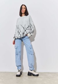 Pulover oversized de culoare gri deschis cu un model abstract negru, purtat cu pantaloni cargo de culoare albastră deschisă, care au buzunare, și încălțăminte chunky în două tonuri.
