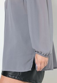 Helles graues Blusenshirt mit langen Ärmeln, gerafften Bündchen und seitlichen Schlitzen, kombiniert mit einem schwarzen Lederrock, der eine glatte Stoffstruktur zeigt.
