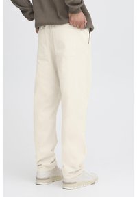 Pantalones color beige claro de corte holgado, con dos bolsillos traseros, combinados con zapatos grises, vistos desde atrás sobre un fondo neutro.