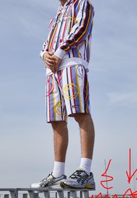 Witte hoodie en shorts met verticale veelkleurige strepen en een lachende gezichtjespatroon. Draag met grijs en zwarte hardloopschoenen en witte sokken.