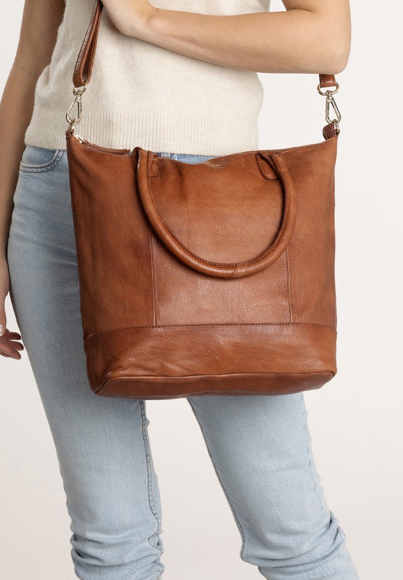 Bolso tote de cuero marrón con asas dobles, cierre de cremallera y una textura suave. Presenta un detalle de costura en la parte inferior y una silueta elegante.