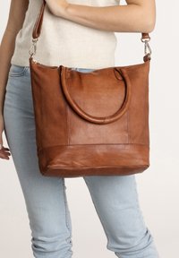 Bruine leren tote bag met dubbele handvatten, ritssluiting en een gladde textuur. Bevat een detail bij de ondernaad en heeft een strakke silhoutte.
