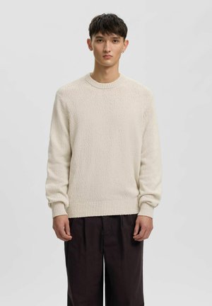 Ung mand med kort mørkt hår iført en cremefarvet tekstureret sweater og løse sorte bukser, stående mod en ensfarvet baggrund.
