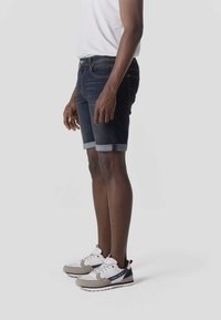 Mężczyzna stojący, ubrany w ciemne, podwinięte szorty dżinsowe, białe trampki z szarymi i czarnymi akcentami oraz białą koszulkę, widok z boku.