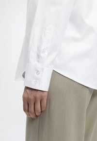 Λευκό πουκάμισο με μακριά μανίκια από λεία ύφανση, με μυτερή μανσέτα και τρία διάφανα κουμπιά. Το πουκάμισο είναι μπακλατζέ μέσα σε ανοιχτό μπεζ παντελόνι.