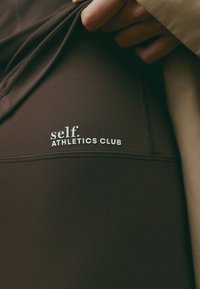 Ruskeat urheilutrikoot, joissa on sileä rakenne ja ommellut yksityiskohdat. Valkoinen "self."-logo "ATHLETICS CLUB" -tekstin yläpuolella lihavoidulla fontilla.