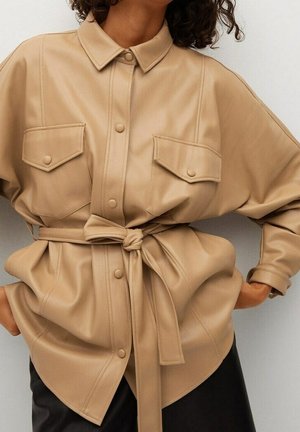Femme portant une chemise ceinturée en simili cuir beige avec poches à rabat boutonnées et manches longues, associée à un pantalon noir.