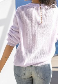 Fioletowy sweter dziany o luźnym kroju, z detalem w postaci guzików z tyłu i ściągaczami na rękawach, w zestawieniu z jasnoniebieskimi dżinsami. Teksturowany materiał.