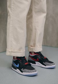 Baskets Nike avec un corps noir, des accents rouges et bleus, et un motif en imprimé éléphant sur la semelle. Associes à un pantalon beige clair retroussé.