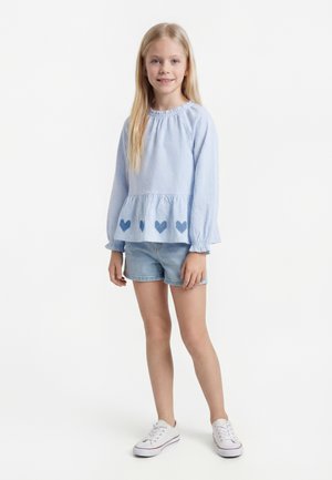 Ung pige, der står, iført en lyseblå langærmet bluse med hjerteprint, denimshorts og hvide sneakers på en hvid baggrund.