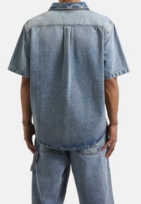 Denimowa koszula z krótkim rękawem z kołnierzykiem, w jasnoniebieskim, wyblakłym odcieniu, z widocznymi detalami szwów na dolnej krawędzi i ramionach.