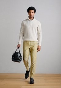 Άσπρο ριγέ πουλόβερ με φερμουάρ, ανοιχτό κίτρινο παντελόνι και μαύρη τσάντα. Μαύρα δερμάτινα παπούτσια. Μοντέλο που περπατά σε ξύλινο πάτωμα με φόντο ανοιχτό γκρι.