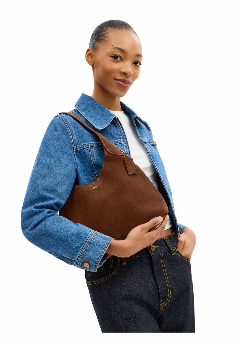 En ung kvinde iført en blå denimjakke og mørke jeans holder en brun ruskind skuldertaske med én hånd i taljehøjde.