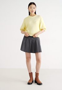 Suéter amarelo de malha com mangas curtas, combinado com uma saia plissada preta e botas de tornozelo castanhas com um acabamento texturizado.
