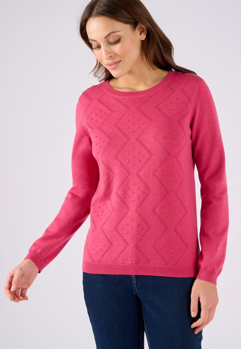 Roze gebreide trui met een ruitpatroon en textuuraccenten, lange mouwen en een ronde halslijn, gecombineerd met donkerblauwe jeans.