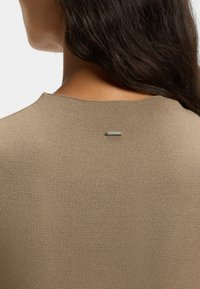 Close-up of a person’s back wearing a beige knit sweater with a small silver rectangular tag near the neckline.
