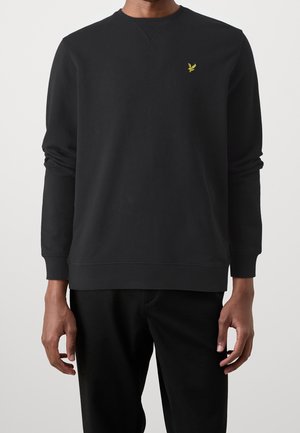 Mann trägt ein schlichtes schwarzes Langarm-Sweatshirt mit einem kleinen gelben Vogel-Logo auf der linken Brust, kombiniert mit einer schwarzen Hose.