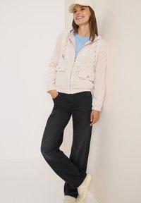 Lichroze corduroy zip-up jas met trekkoordcapuchon, twee voorkwestjes, gedragen over een blauwe shirt en zwarte broek, gecombineerd met lichte schoenen.
