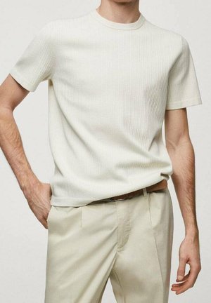 Mann trägt ein cremefarbenes, geripptes Kurzarmshirt, das in beige Hosen mit braunem Gürtel gesteckt ist, steht mit einer Hand in der Tasche.