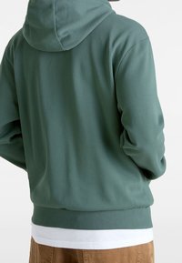 Sudadera con capucha verde con tejido texturizado, capucha y puños acanalados. Se lleva sobre una camisa blanca, combinada con pantalones marrones. Diseño sencillo, sin estampados.