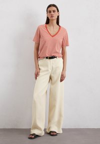 Gestreiftes orange-weißes V-Ausschnitt-T-Shirt, kombiniert mit weiten cremefarbenen Hosen und schwarzen Sandalen. Einfaches Design mit lockerer Passform und strukturiertem Stoff.
