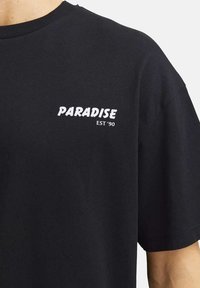 T-shirt nera in cotone con la parola "PARADISE" in carattere bianco stilizzato e "EST '90" sotto di essa. Design a maniche corte e colletto rotondo.