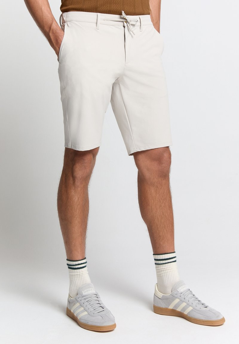 Beige shorts met een trekkoord in de taille, gemaakt van lichtgewicht materiaal, gecombineerd met grijze sportschoenen en witte sokken met groene strepen.