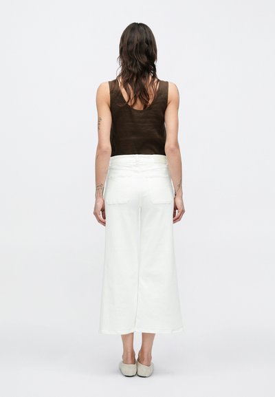 Καφέ αμάνικη μπλούζα με στρογγυλή λαιμόκοψη σε συνδυασμό με λευκό ψηλόμεσο παντελόνι με φαρδιά μπατζάκια. Το ύφασμα φαίνεται απαλό με ελαφρώς υφή.