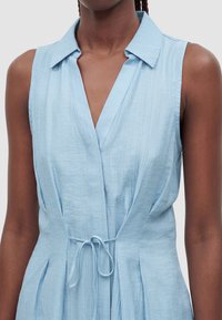 Robe sans manches bleu clair en tissu texturé avec col, présentant un décolleté en V et un détail de ceinture à la taille sur une personne à la peau foncée.