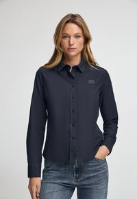 Camicia button-up blu scuro in un tessuto liscio, dotata di colletto, maniche lunghe e una piccola etichetta con marchio sul lato sinistro del petto.