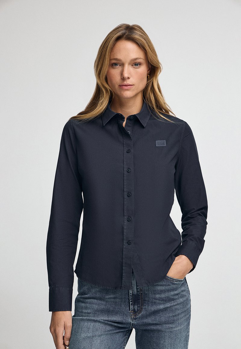 Camicia button-up blu scuro in un tessuto liscio, dotata di colletto, maniche lunghe e una piccola etichetta con marchio sul lato sinistro del petto.