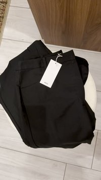 Pantaloni negri fabricați dintr-un material neted, cu buzunare laterale și o etichetă atașată. Pliate cu grijă pe un scaun de culoare deschisă.