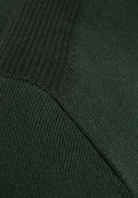 Grüne Pulloverstoff mit unterschiedlichen Texturen; glatte Bereiche, die von gerippten Mustern kontrastiert werden und ein gemischtes Materialbild zeigen.