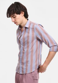 Camisa de botones a rayas en azul claro y rosa suave, que presenta líneas horizontales texturizadas y un collar clásico. Las mangas dobladas añaden un detalle.